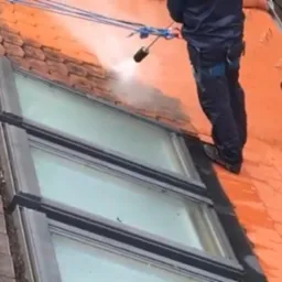 Pracownik w kombinezonie czyści dachówkę wokół okna dachowego za pomocą myjki ciśnieniowej, widoczny efekt czyszczenia między starą a nową powierzchnią.