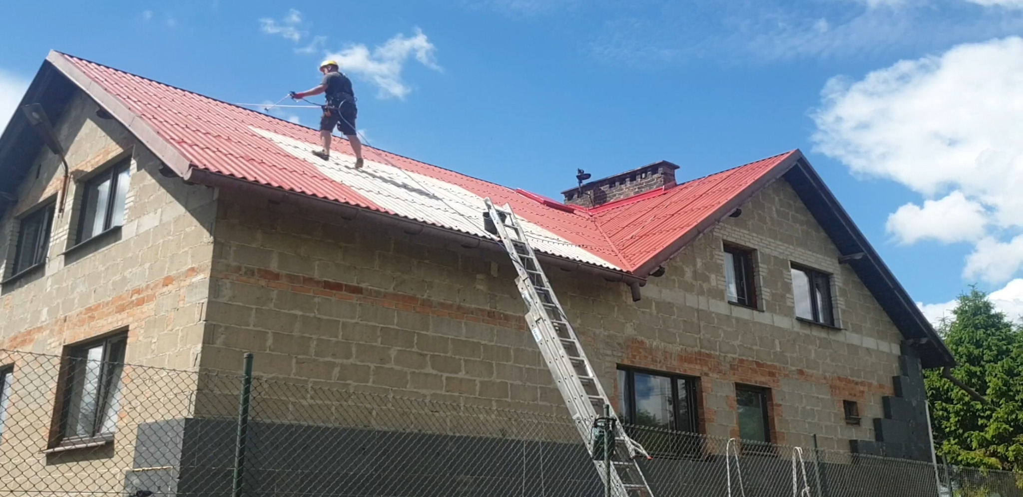 Pracownik w uprzęży, z lancą natryskową, częściowo umyty dach z blachy falistej na dwukondygnacyjnym domu z szarego bloczka, drabina oparta o dach, błękitne niebo.