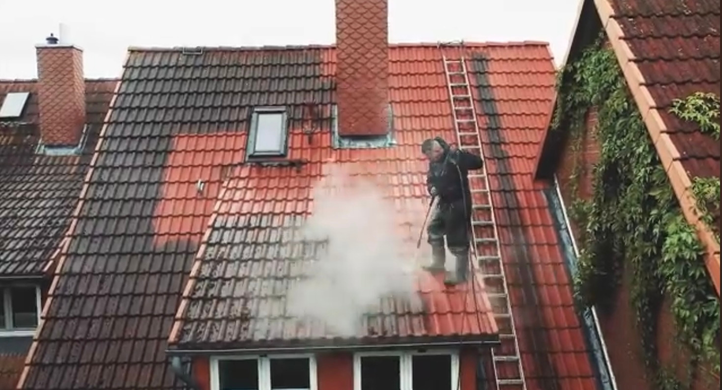 Pracownik w kombinezonie i gumowych butach czyści czerwoną dachówkę myjką ciśnieniową, widoczna różnica między umytą a brudną częścią dachu, obok drabina i porośnięta bluszczem ściana budynku.