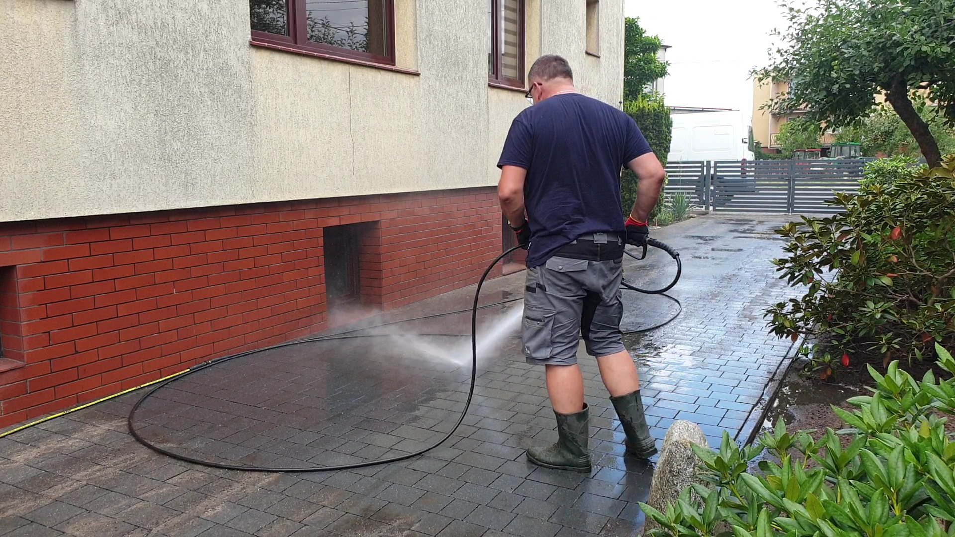 Mężczyzna w gumowych butach czyści kostkę brukową myjką ciśnieniową w pobliżu ceglanej ściany budynku.