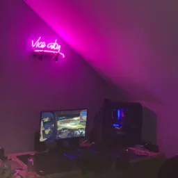 Stanowisko komputerowe w pokoju na poddaszu, oświetlone neonem z napisem 'Vice City', monitor wyświetla grę, obudowa komputera z podświetleniem LED, klawiatura i mysz na biurku, dodatkowy stolik...