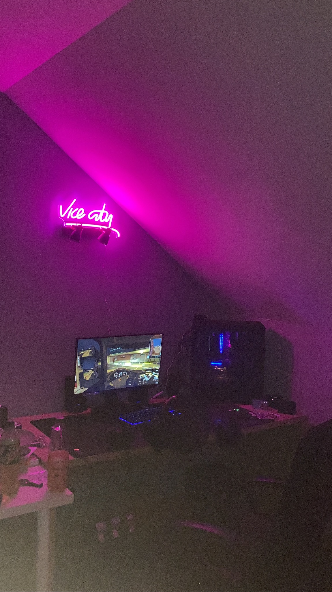 Stanowisko komputerowe w pokoju na poddaszu, oświetlone neonem z napisem 'Vice City', monitor wyświetla grę, obudowa komputera z podświetleniem LED, klawiatura i mysz na biurku, dodatkowy stolik...