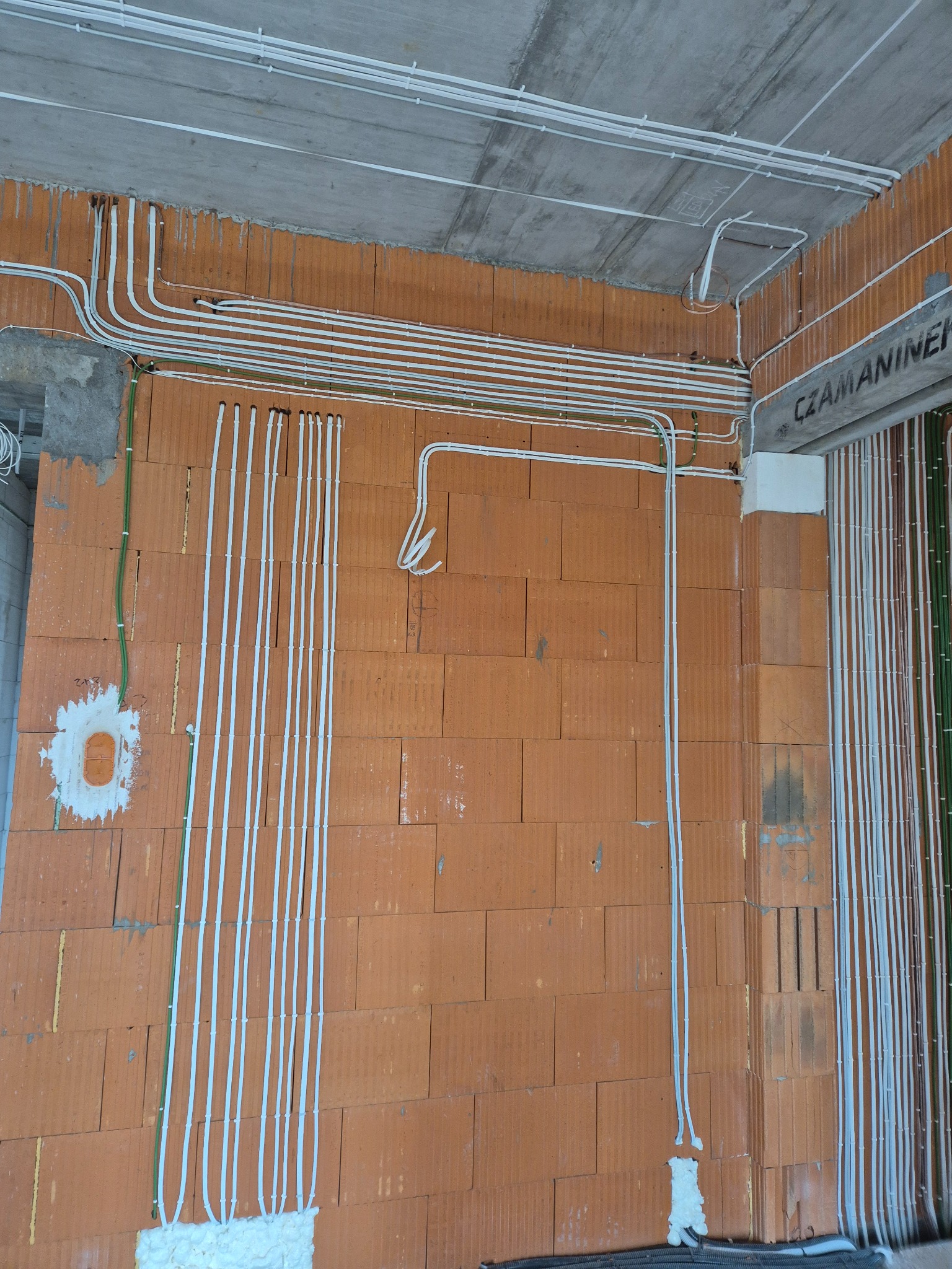 Białe rurki instalacji elektrycznej prowadzone po ceglanej ścianie i betonowym suficie w surowym stanie. Widoczne kable i pianka montażowa.