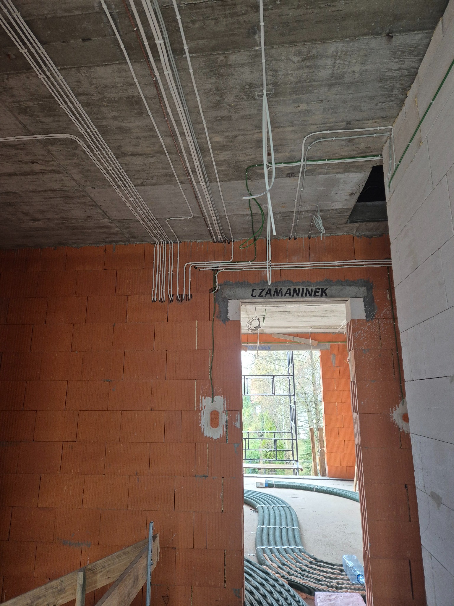 Wnętrze w trakcie budowy z widocznymi przewodami elektrycznymi ułożonymi na suficie i ścianach, prowadzącymi do otworu drzwiowego. Widoczne kable w peszlach na podłodze.