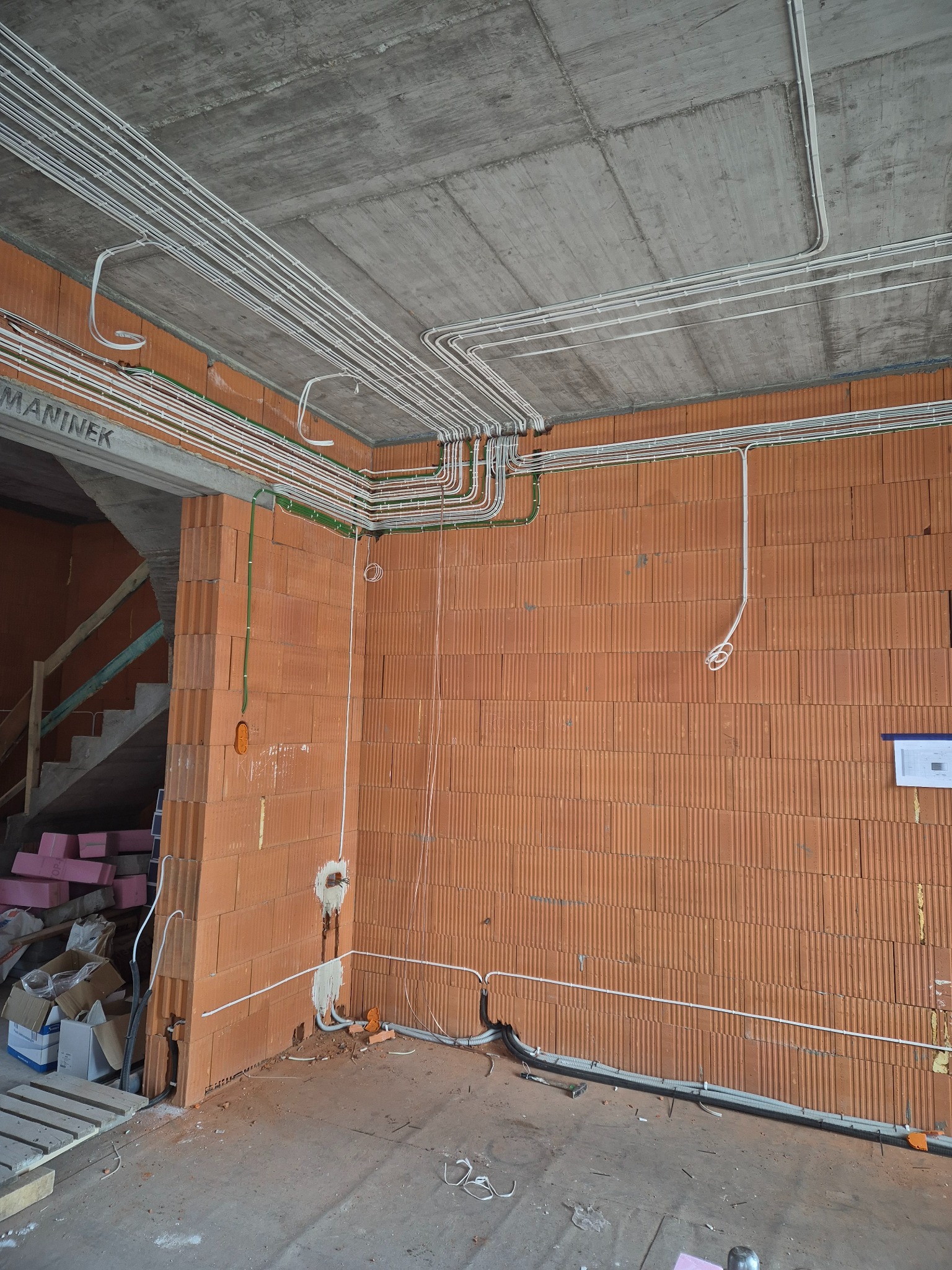 Instalacja elektryczna w surowym budynku: białe kable prowadzone po suficie i ścianach z cegły, widoczne puszki instalacyjne i fragment schodów. Stan surowy.