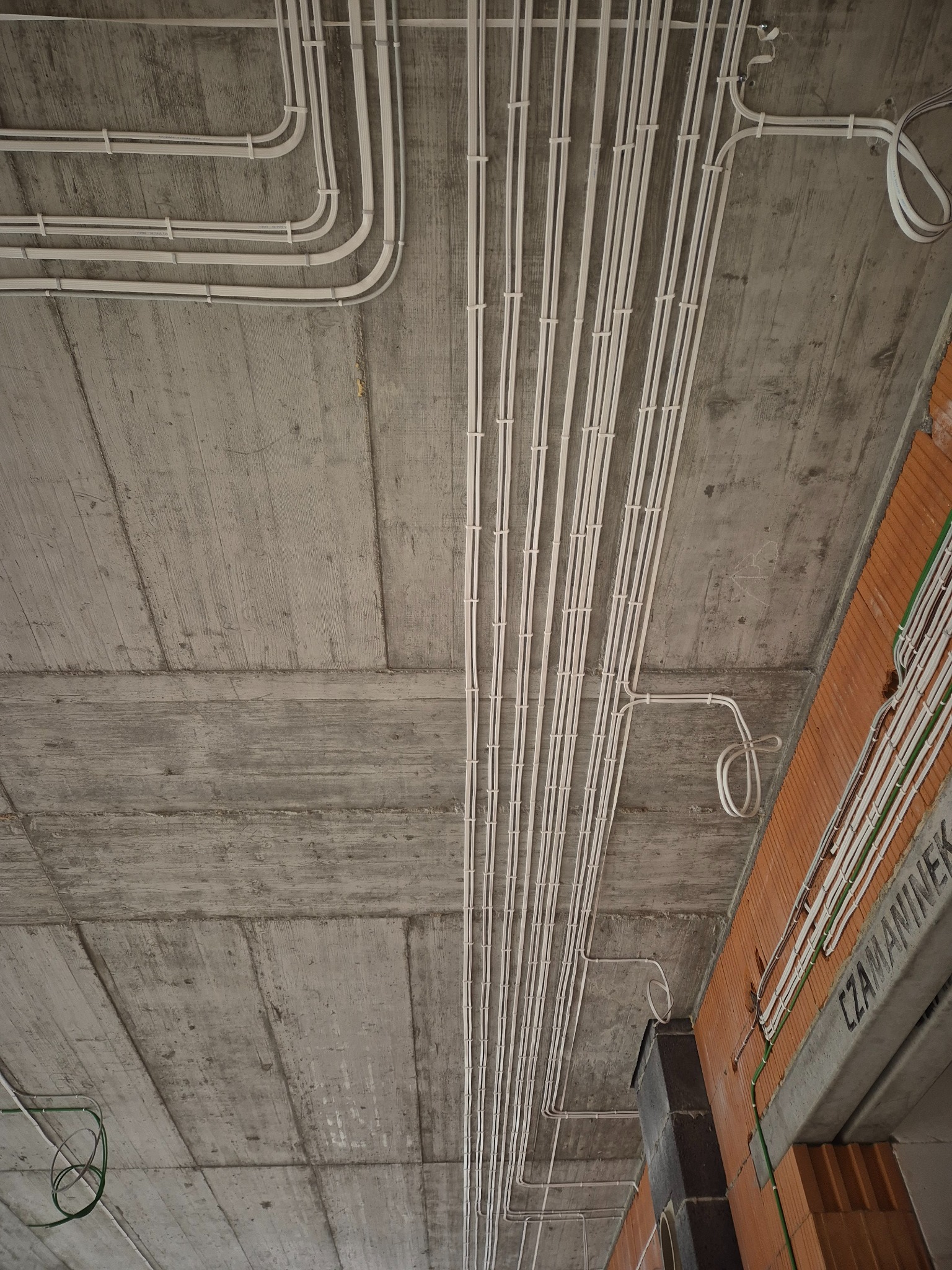 Betonowy strop z równolegle ułożonymi, białymi rurami instalacji elektrycznej, przymocowanymi uchwytami. Widoczne fragmenty muru z cegły dziurawki.