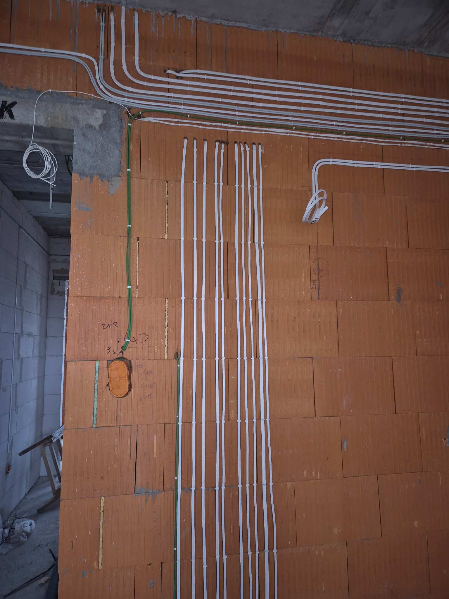 Białe i zielone kable elektryczne prowadzone po ceglanej ścianie w surowym stanie, widoczne oznaczenia i puszka instalacyjna. Etap prac instalacyjnych w budynku.