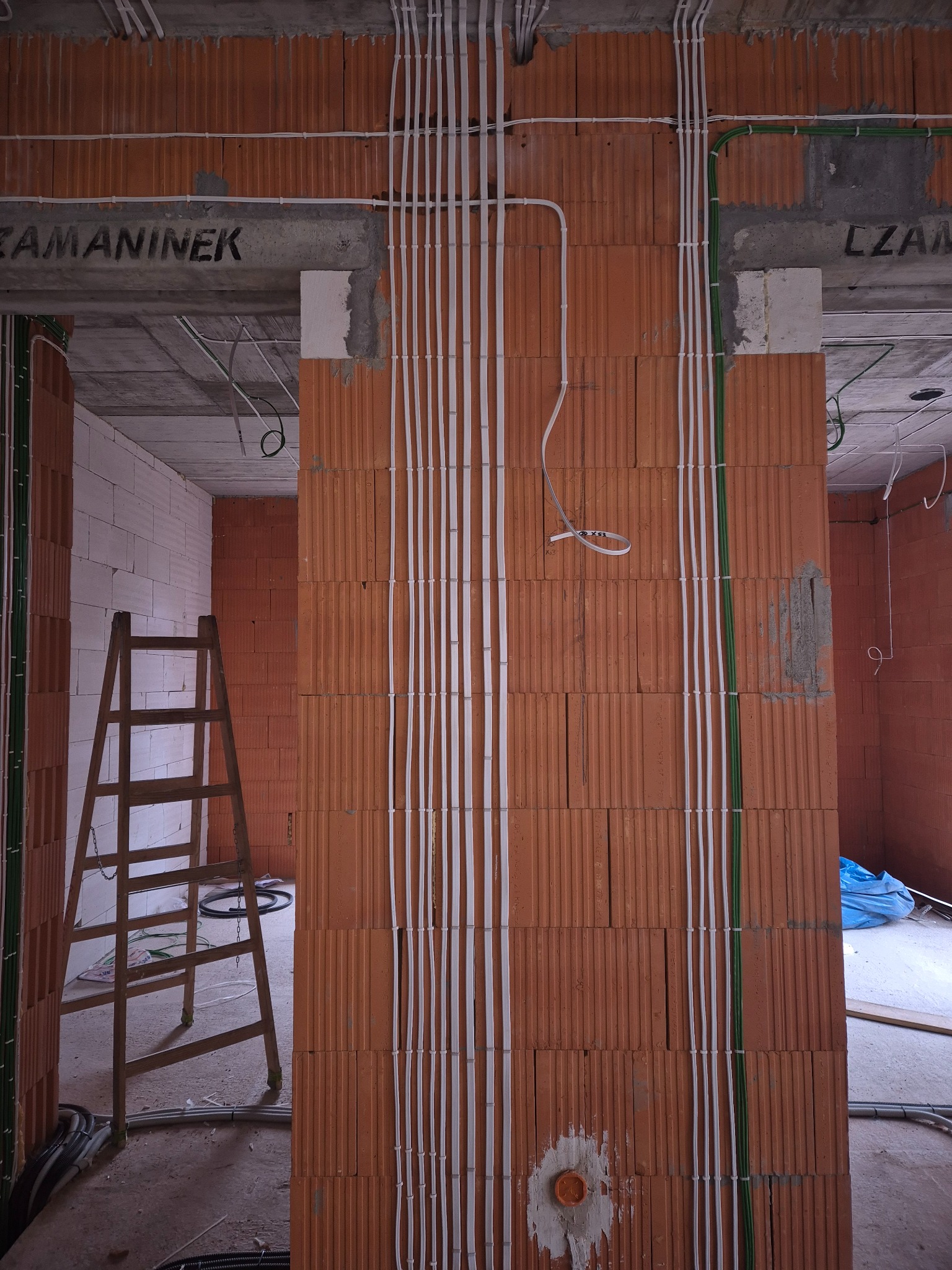 Wewnętrzna instalacja elektryczna w budynku w trakcie budowy. Białe i zielone kable prowadzone pionowo po ceglanej ścianie. Widoczna drabina i fragmenty innych pomieszczeń.