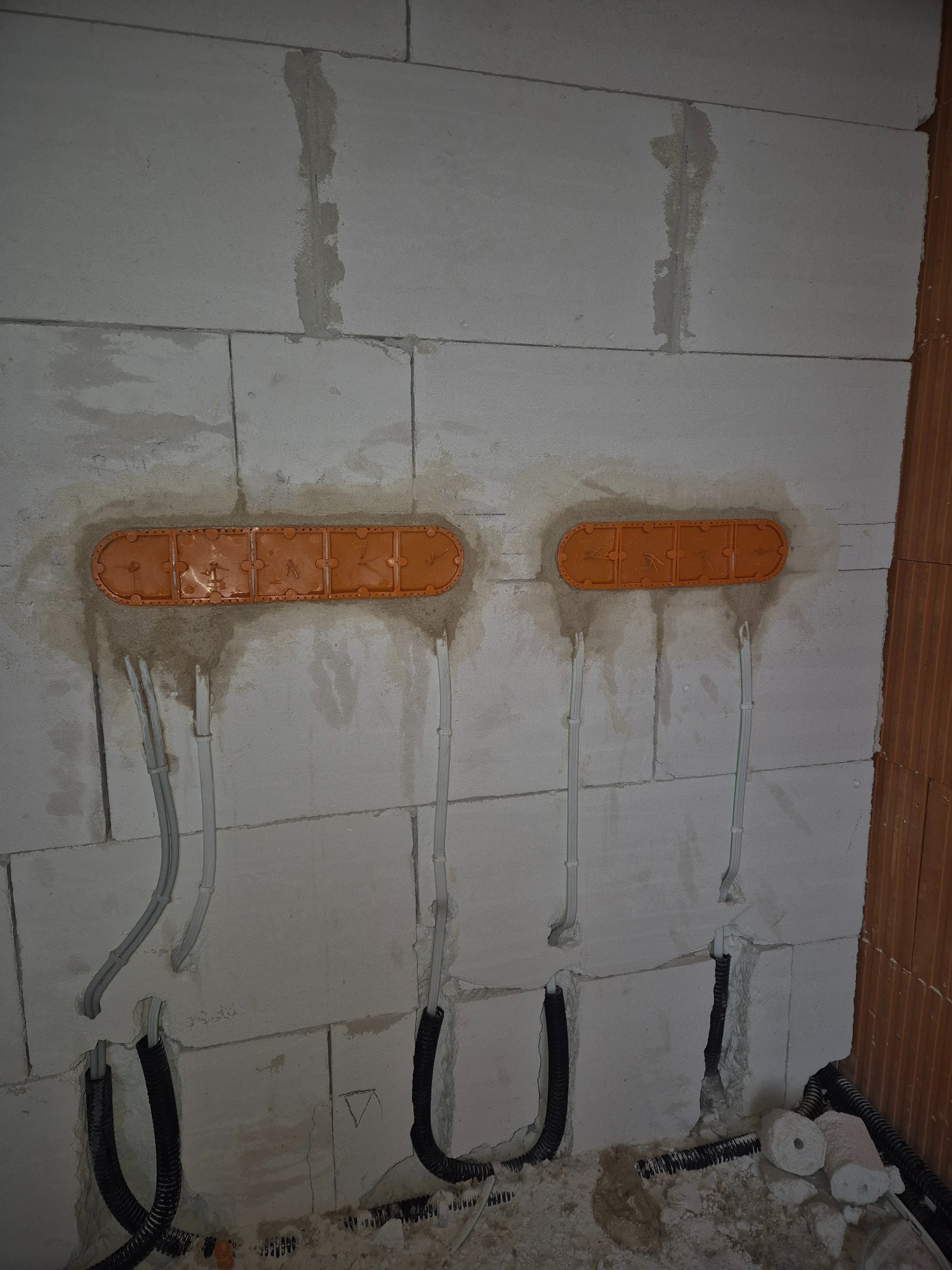 Dwa rzędy pomarańczowych puszek instalacyjnych wmurowane w białą ścianę z betonu komórkowego, z widocznymi przewodami elektrycznymi w peszlach i bez.