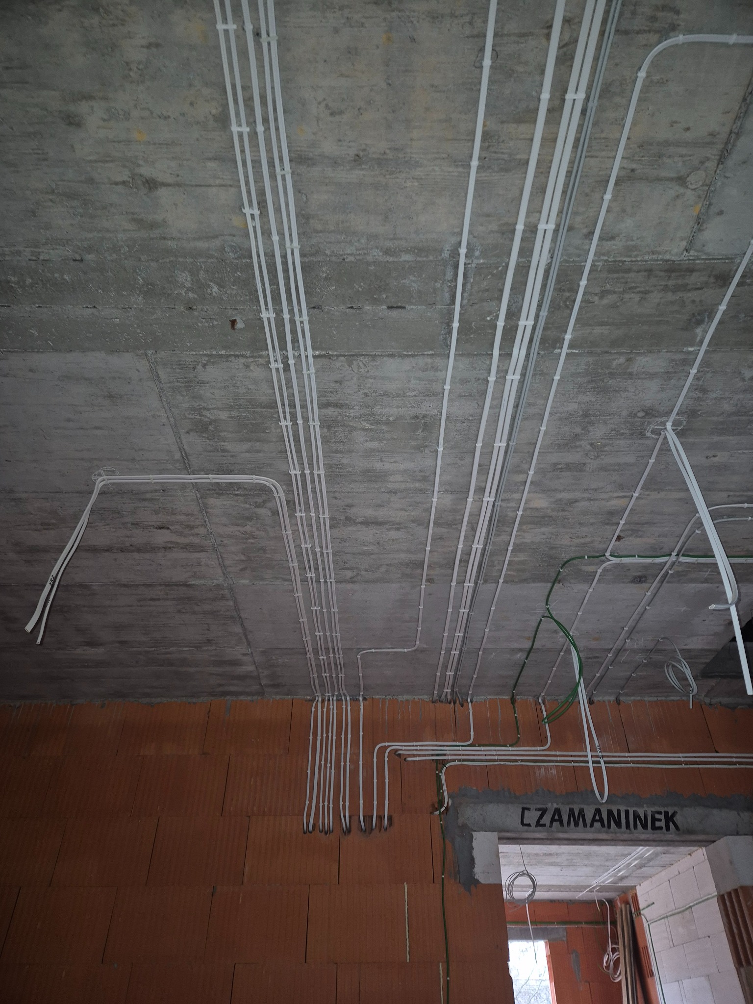 Białe kable elektryczne przymocowane do surowego betonowego sufitu w nowym budynku z ceglanymi ścianami. Widoczne przewody w peszlach i nadproże z napisem.