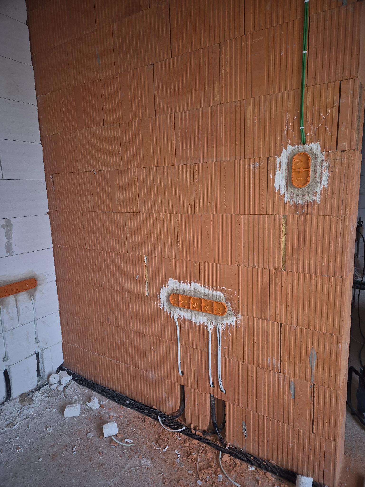Ceglana ściana z zamontowanymi puszkami elektrycznymi i przewodami w trakcie instalacji. Widoczne kable i peszle. Surowy stan, instalacja elektryczna w trakcie realizacji.