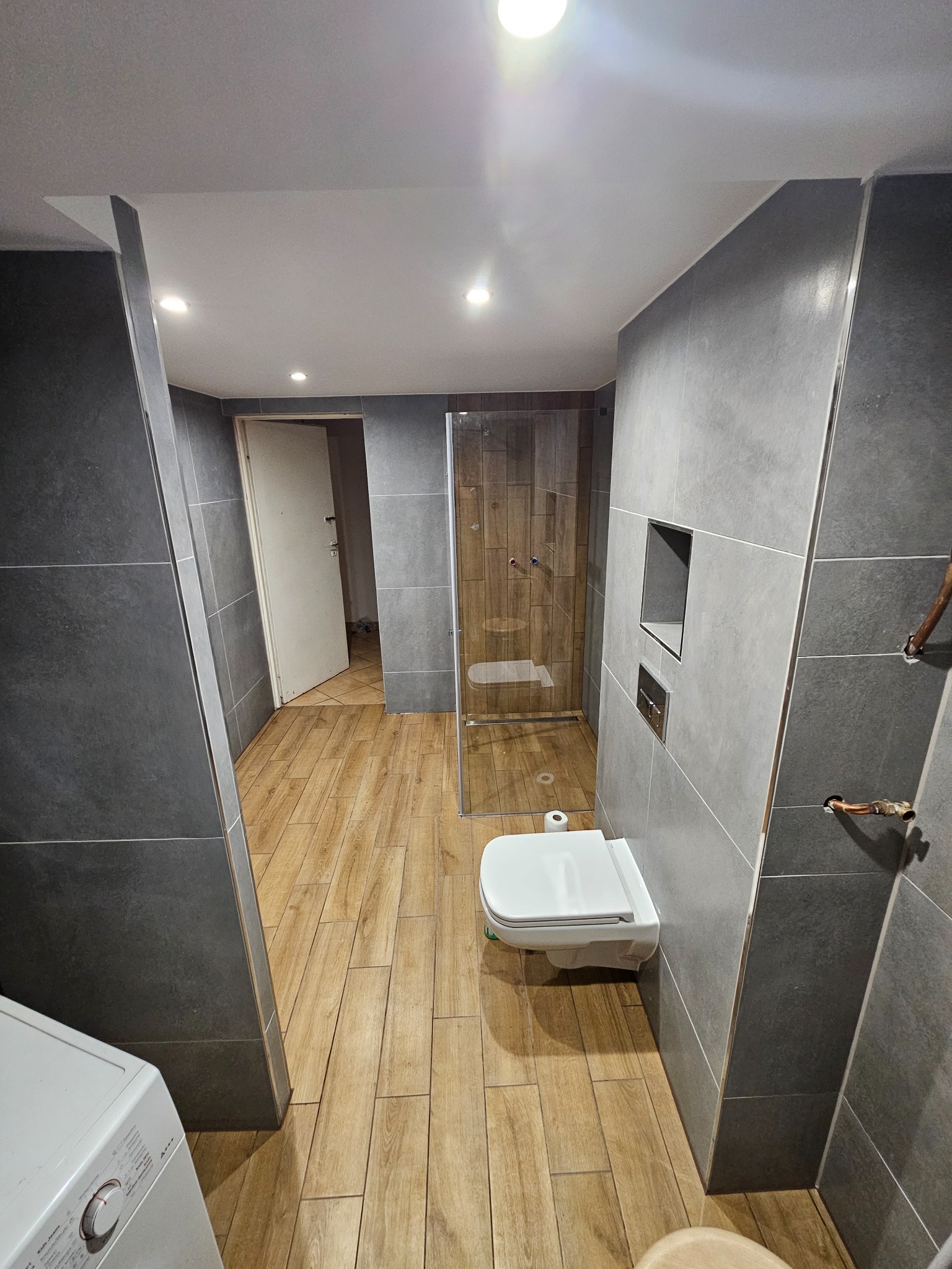 Nowoczesna łazienka w Nasielsku z szarymi płytkami, drewnopodobną podłogą, kabiną prysznicową i podtynkową toaletą, widziana z perspektywy wejścia.