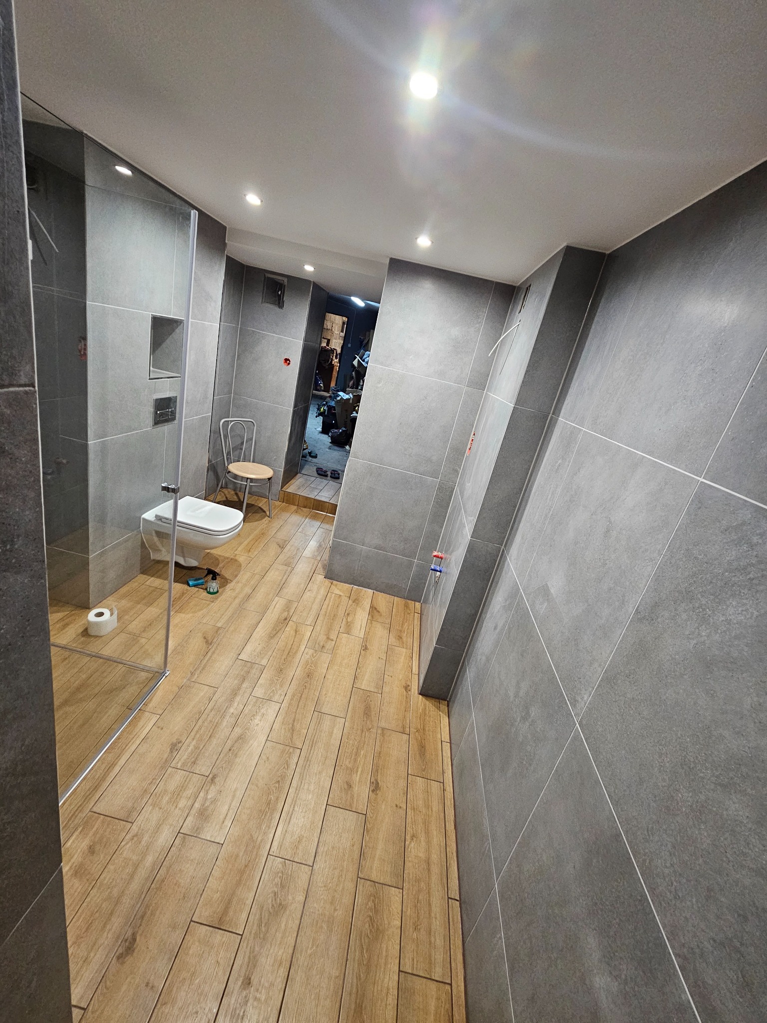 Nowoczesna łazienka z szarymi płytkami na ścianach i drewnopodobną podłogą, widoczna kabina prysznicowa ze szklaną ścianką, toaleta podwieszana i krzesło z drewnianym siedziskiem w tle.