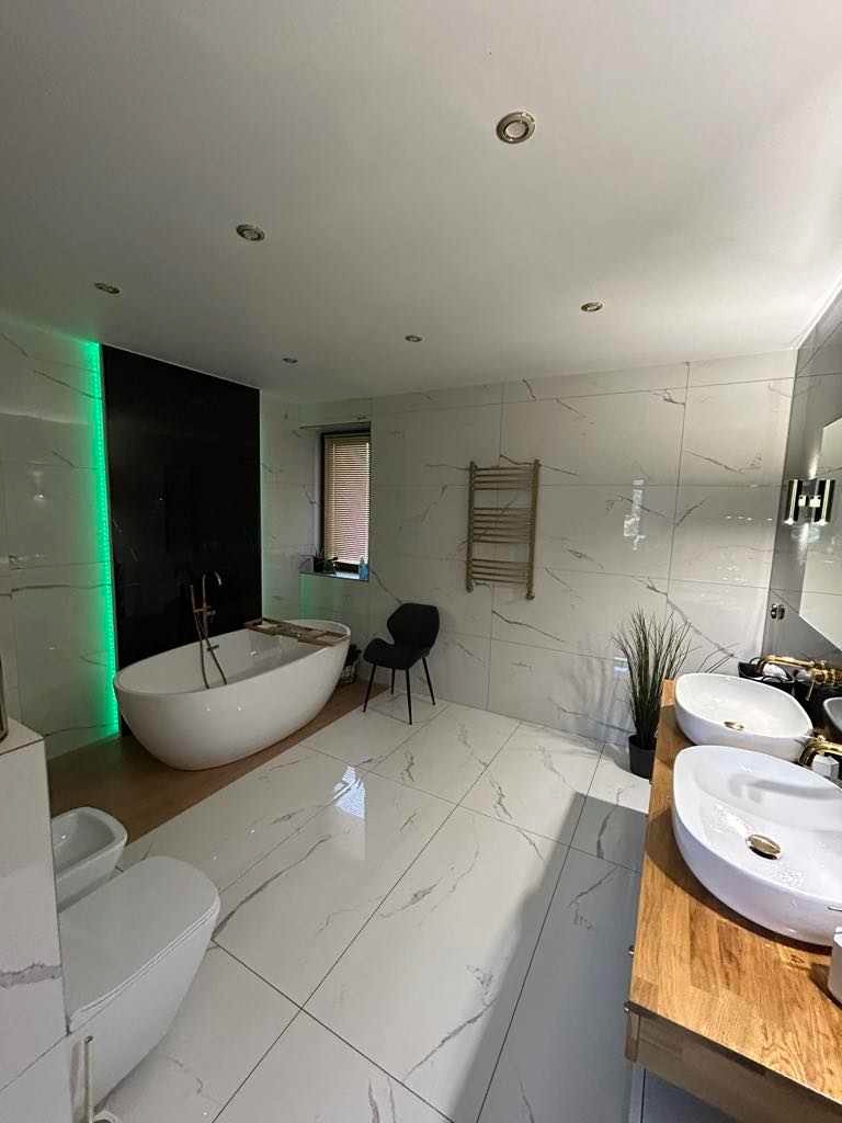 Nowoczesna łazienka z wolnostojącą wanną na drewnianym podeście, podświetleniem LED w kolorze zielonym, białymi płytkami imitującymi marmur, dwoma umywalkami nablatowymi na drewnianym blacie...