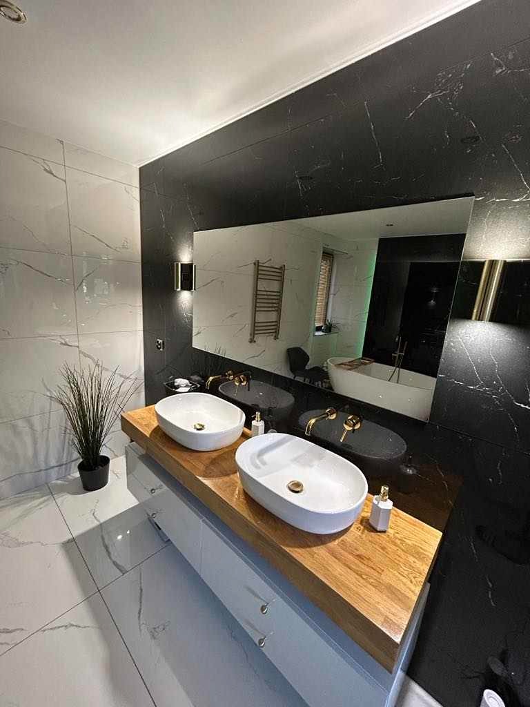 Nowoczesna łazienka z dwoma białymi umywalkami nablatowymi na drewnianym blacie, szare płytki imitujące marmur na ścianach, duże lustro odbijające wannę i grzejnik drabinkowy, oświetlenie punktowe.