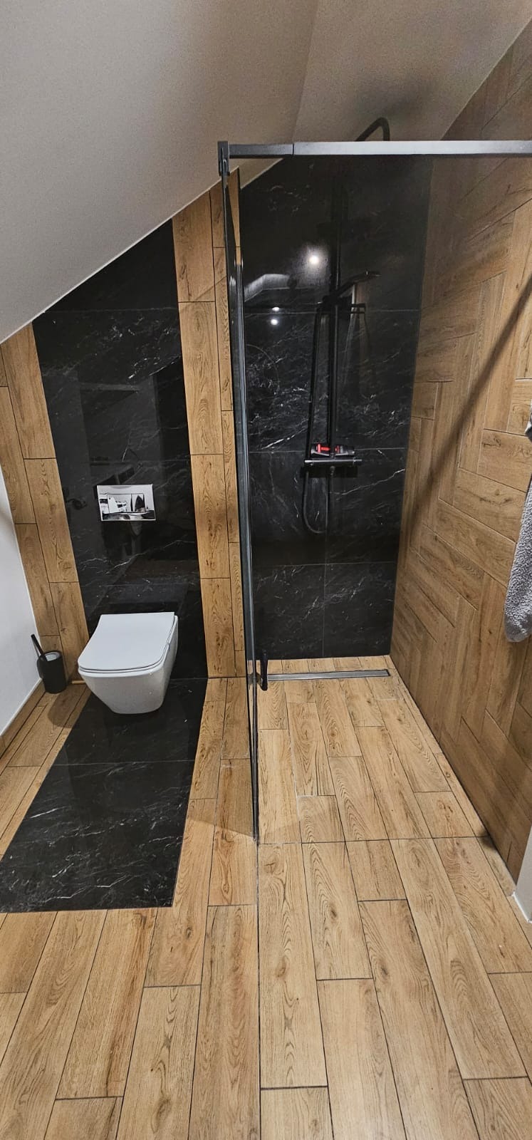 Nowoczesna łazienka na poddaszu z czarnymi marmurowymi płytkami i drewnopodobną podłogą, kabina prysznicowa z czarnymi profilami i wisząca toaleta.