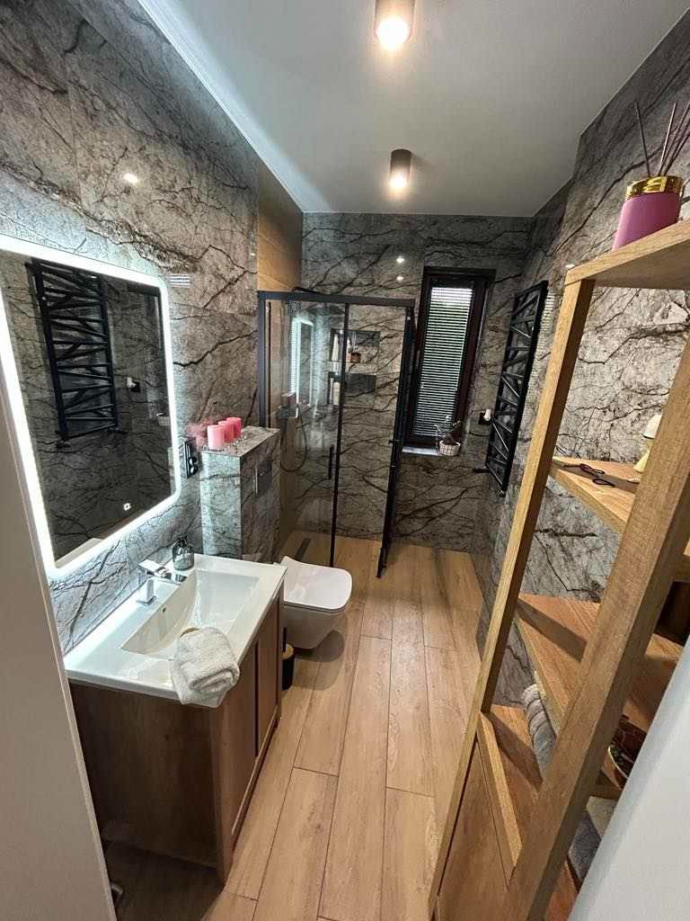 Nowoczesna łazienka z marmurowymi ścianami, drewnopodobną podłogą, kabiną prysznicową ze szkła i czarnymi ramami, podświetlanym lustrem nad umywalką i drewnianym regałem. Dekoracyjne świece...