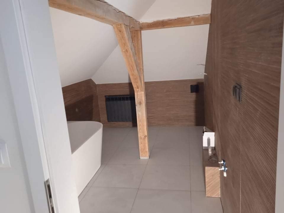 Przestronna łazienka na poddaszu z drewnianą belką konstrukcyjną, płytkami podłogowymi w odcieniach szarości i zabudową z drewnopodobnych paneli na ścianach. Widoczna wanna wolnostojąca i grzejnik...