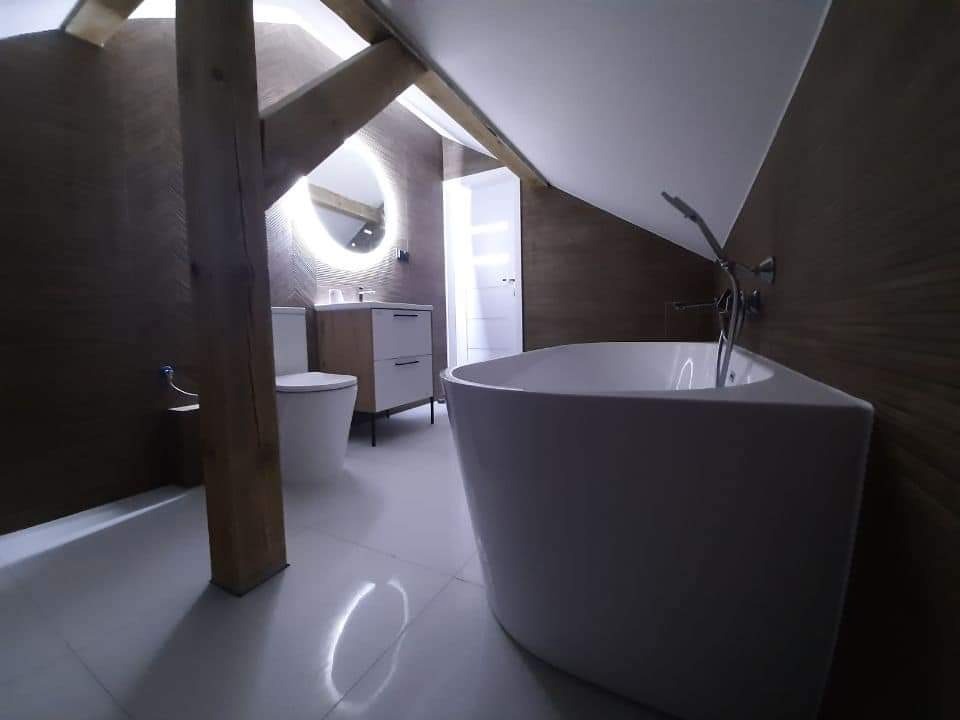 Nowoczesna łazienka na poddaszu z drewnianą belką, okrągłym lustrem z podświetleniem LED, białą wanną narożną i minimalistyczną szafką pod umywalkę.