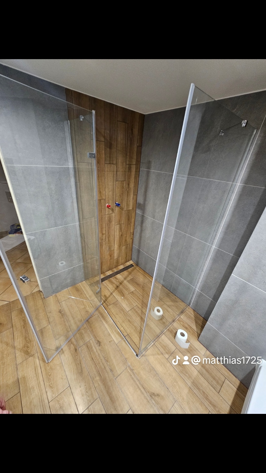 Nowoczesna kabina prysznicowa walk-in z drewnopodobną ścianą i szarymi płytkami, widoczne zawiasy i odpływ liniowy, z rolką papieru toaletowego na podłodze.