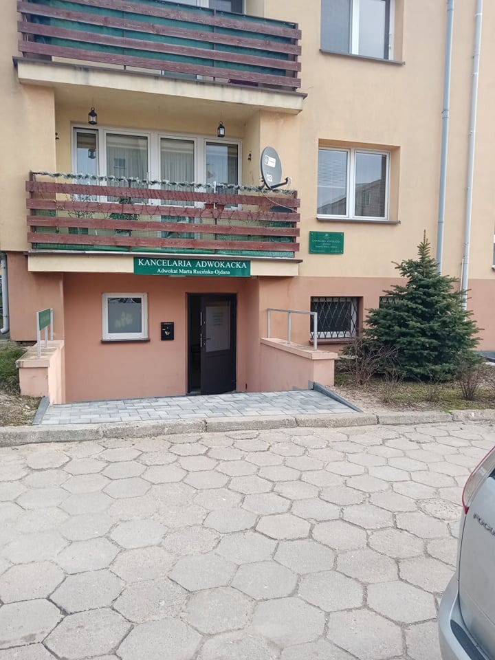Wejście do Kancelarii Adwokackiej z zielonym szyldem informacyjnym, mała choinka obok, balkon z drewnianą balustradą nad wejściem.