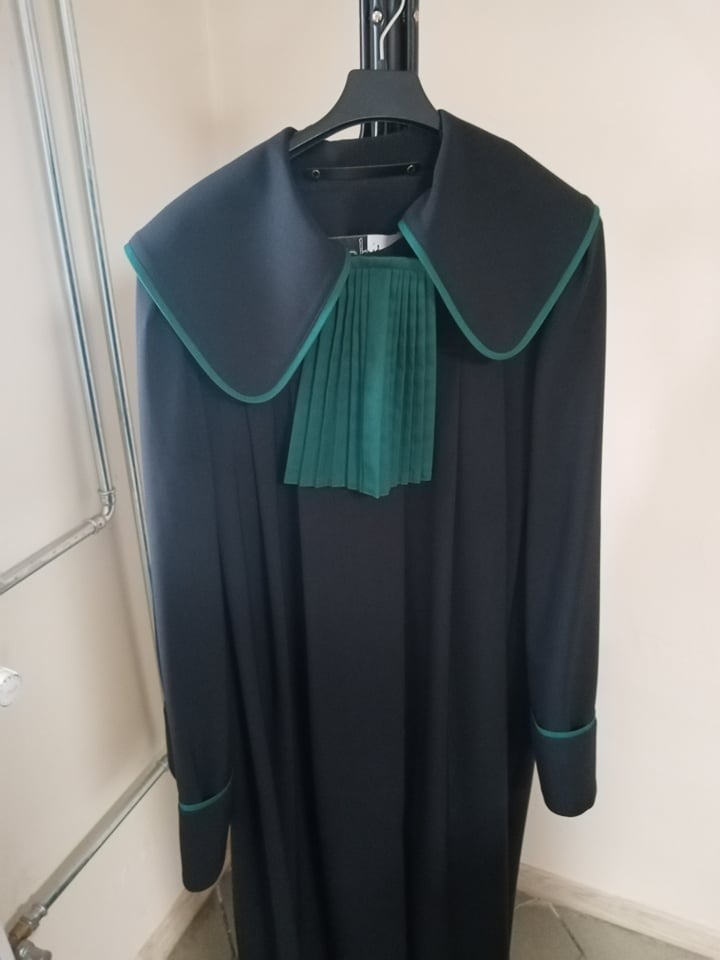 Toga adwokacka zawieszona na wieszaku, czarna z zielonymi elementami, widoczne mankiety i plisowana żabot