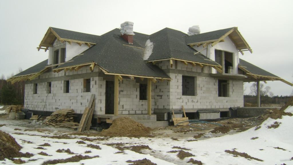 Dwukondygnacyjny dom w stanie surowym zamkniętym, z czarnym dachem pokrytym papą, białe ściany z betonu komórkowego, drewniane elementy konstrukcyjne dachu, na zewnątrz resztki śniegu i stosy...