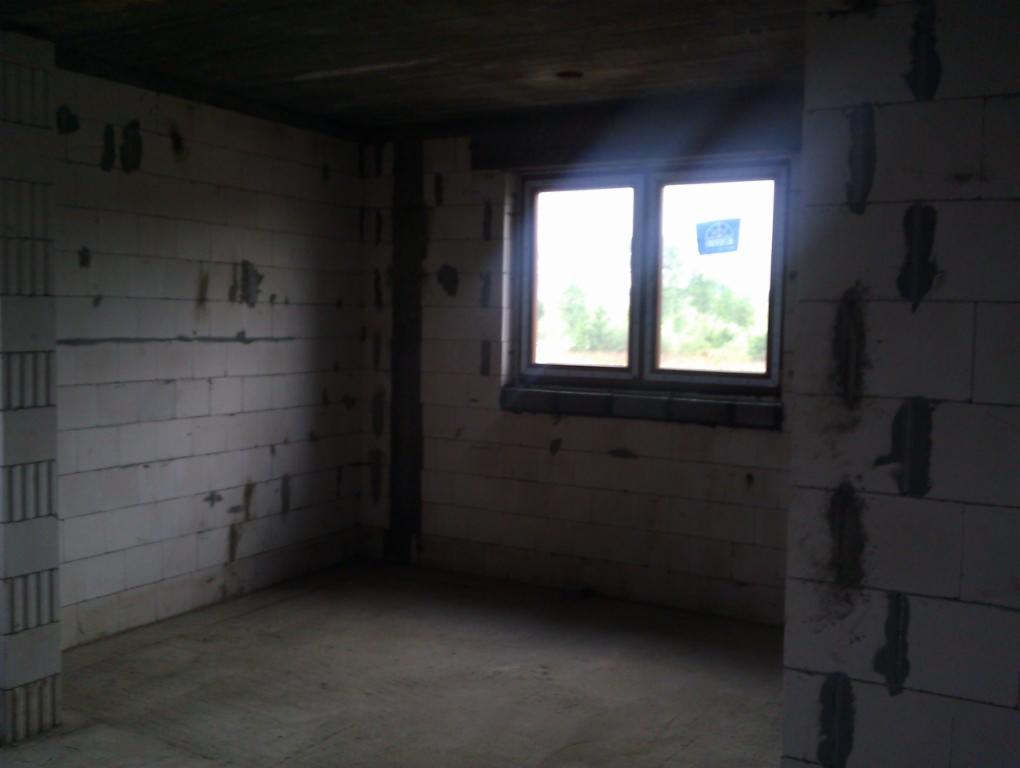 Wnętrze w stanie surowym otwartym, ściany z bloczków, widoczne okno z widokiem na drzewa, betonowa podłoga, sufit w trakcie wykończenia, słabe oświetlenie.