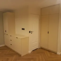 Jasna zabudowa meblowa z szafkami i szufladami, dopasowana do wnęki, z ciemnymi, minimalistycznymi uchwytami, na parkiecie ułożonym w jodełkę.