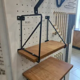 Ekspozycja dwóch drewnianych półek wiszących na metalowym stelażu, przymocowanych do perforowanej ściany z ekspozycją uchwytów meblowych w kolorze złotym i czarnym.