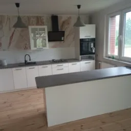 Jasna kuchnia z wyspą, białymi frontami szafek i szarym blatem, wzorzysta tapeta imitująca marmur na ścianie, drewniana podłoga