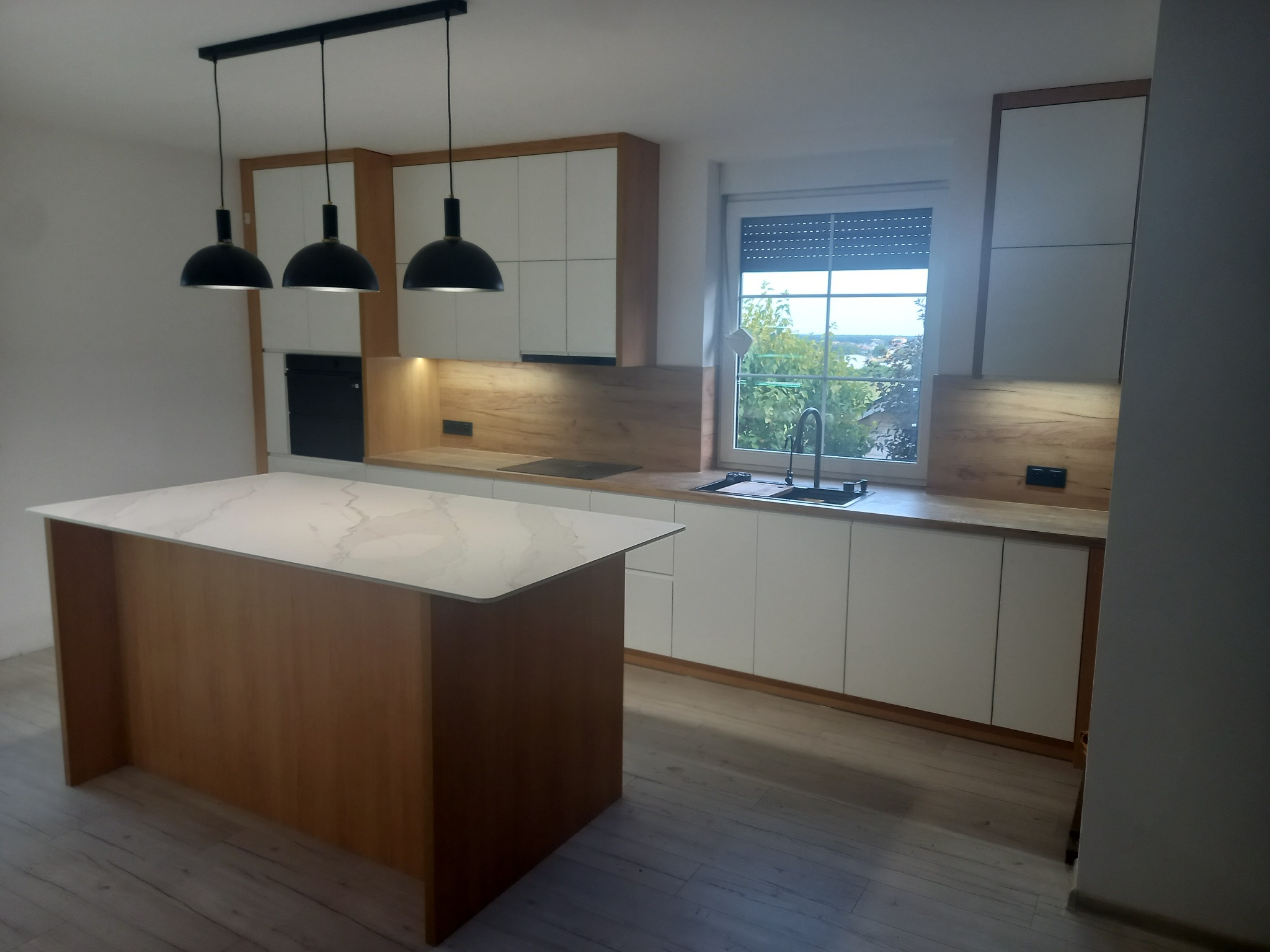 Nowoczesna kuchnia z wyspą: białe szafki z drewnianymi akcentami, blat z marmurowym wzorem na wyspie, trzy czarne lampy wiszące nad wyspą, okno z widokiem na zieleń.