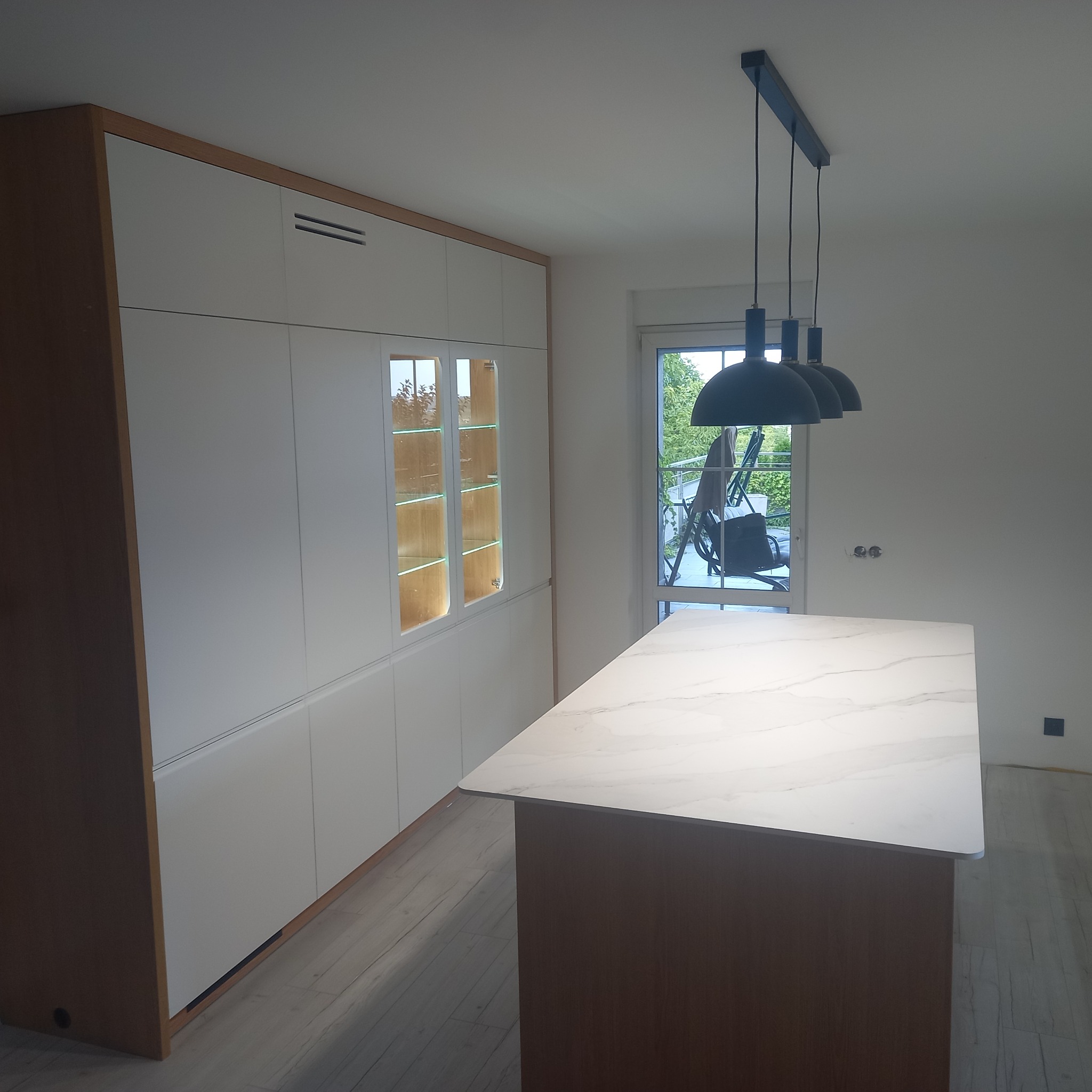 Nowoczesna kuchnia z białymi szafkami z drewnianymi akcentami i wyspą z marmurowym blatem, oświetlona trzema czarnymi lampami wiszącymi.