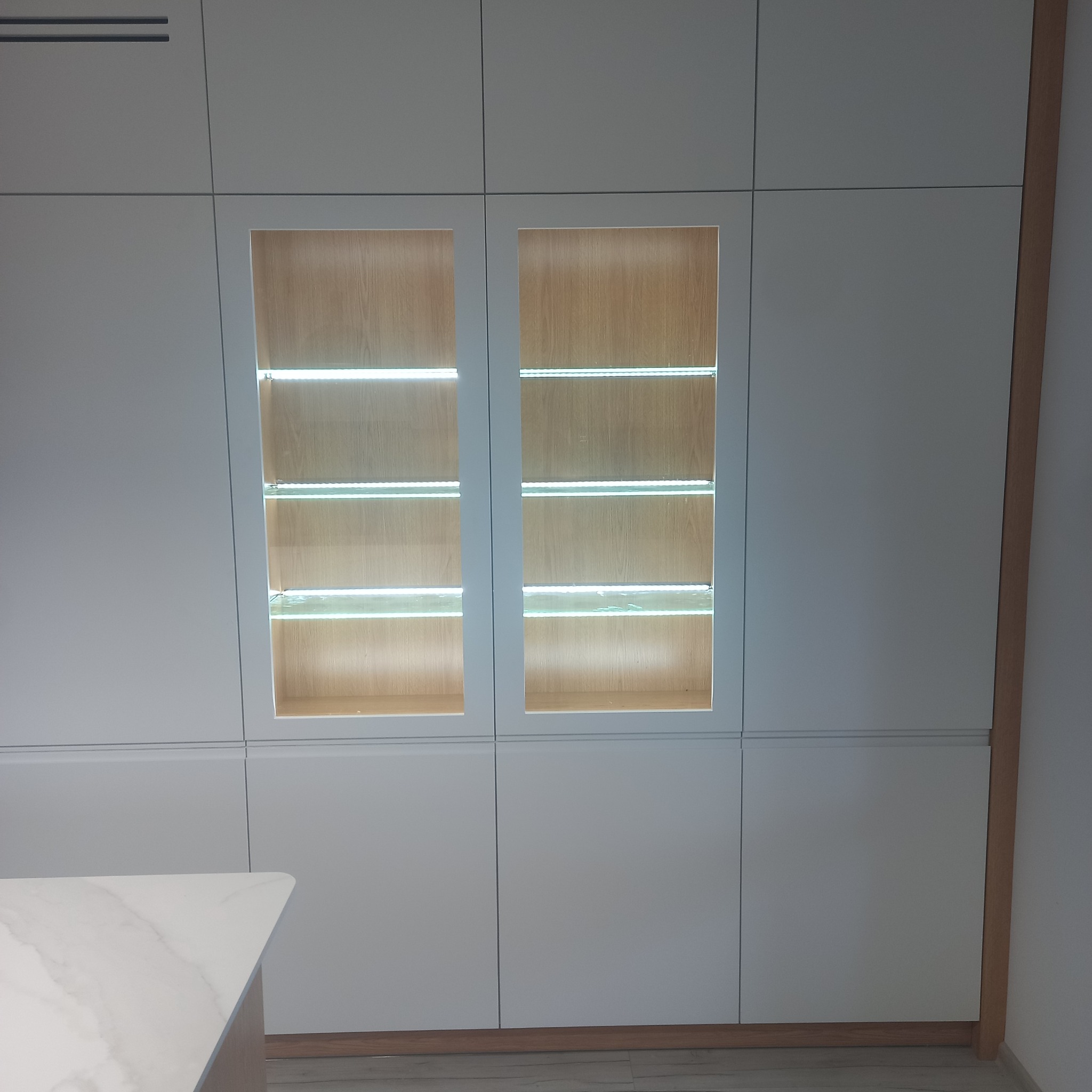 Biała zabudowa kuchenna z podświetlanymi półkami wewnątrz przeszklonych szafek, widoczny blat kuchenny z marmurowym wzorem.