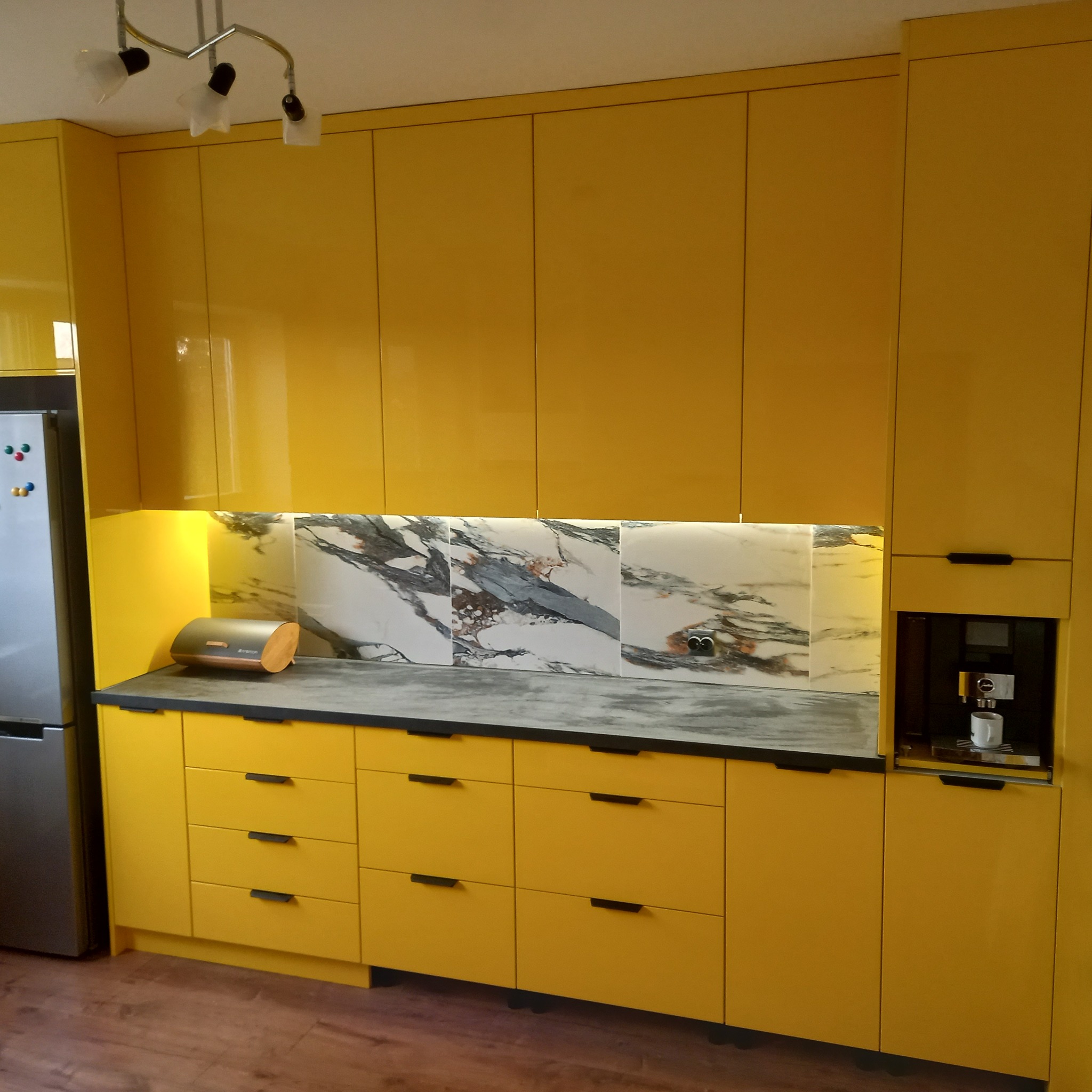 Żółta, lakierowana zabudowa kuchenna z czarnym blatem, marmurowym panelem nad blatem i wbudowanym ekspresem do kawy.