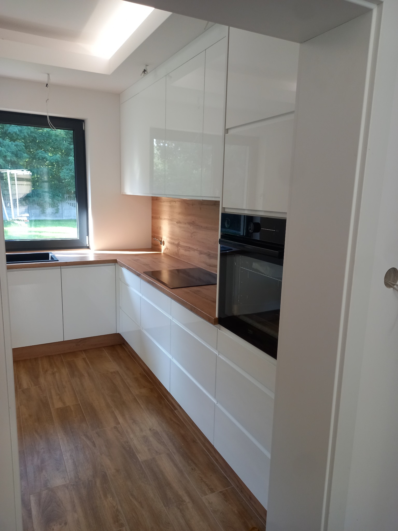 Nowoczesna, biała kuchnia z drewnianym blatem i podłogą, widok z wejścia, czarne okno z widokiem na zieleń.