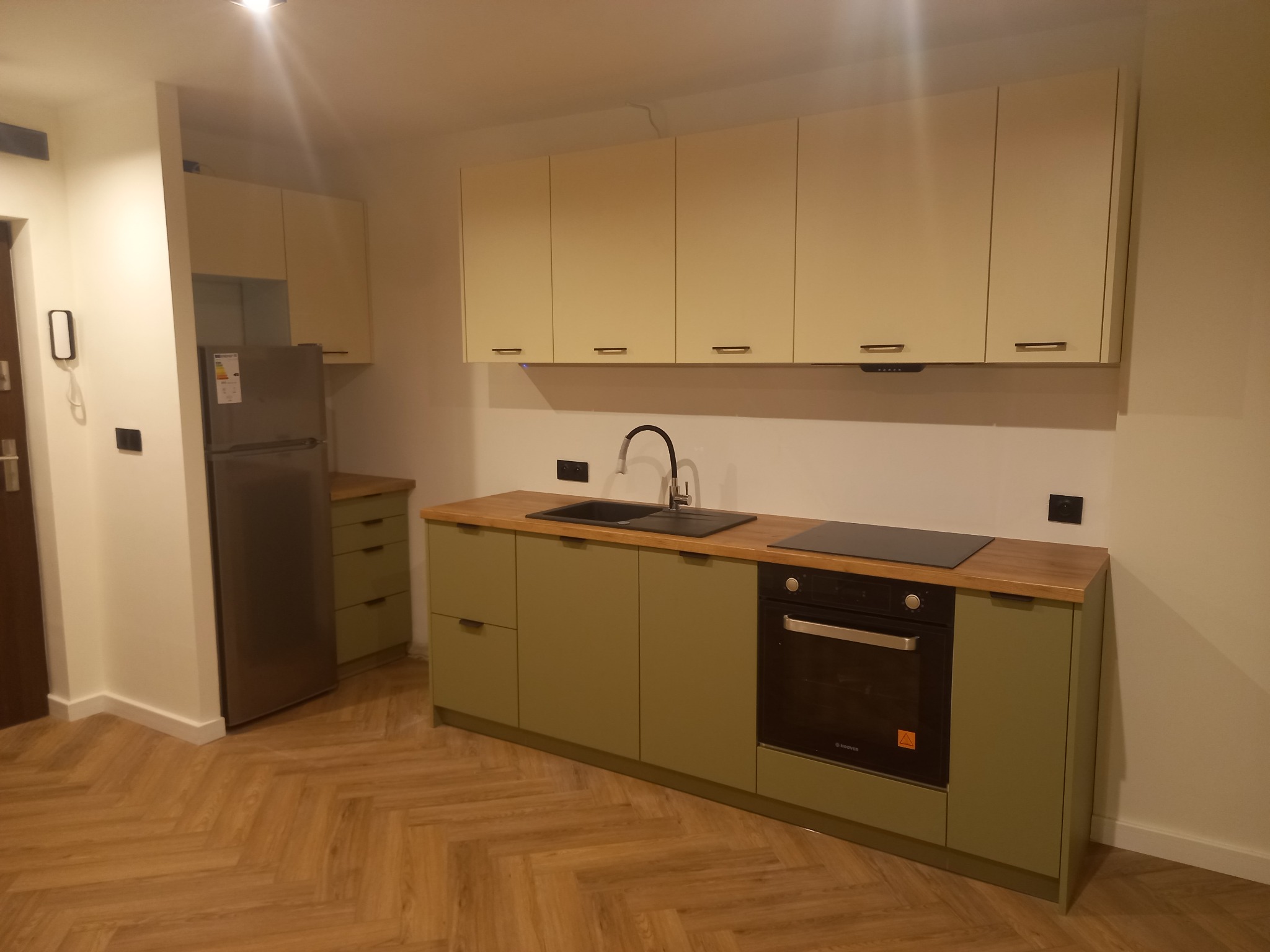 Nowoczesna kuchnia z zielonymi szafkami dolnymi i beżowymi górnymi, drewniany blat, podłoga ułożona w jodełkę, widoczny piekarnik i płyta indukcyjna.