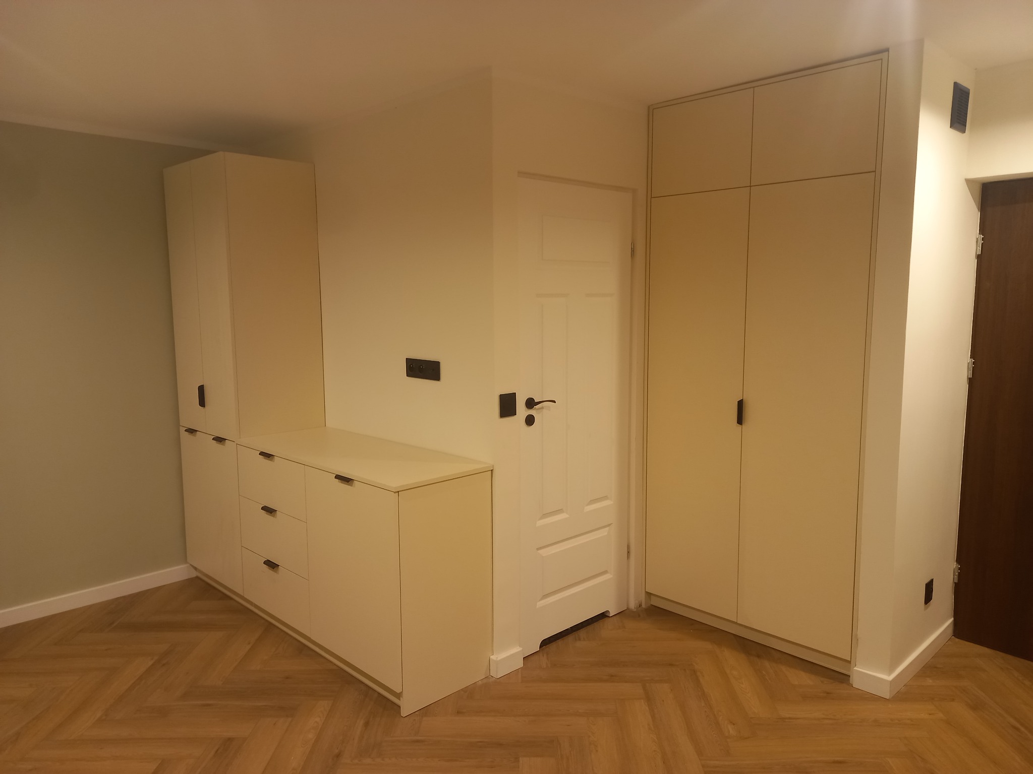 Jasna zabudowa meblowa z szafkami i szufladami, dopasowana do wnęki, z ciemnymi, minimalistycznymi uchwytami, na parkiecie ułożonym w jodełkę.