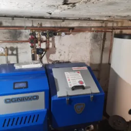 Kotłownia z dwoma niebieskimi kotłami marki Ogniwo i Kospel oraz instalacją hydrauliczną z miedzianych rur na tle surowej ściany z widocznym betonowym stropem.