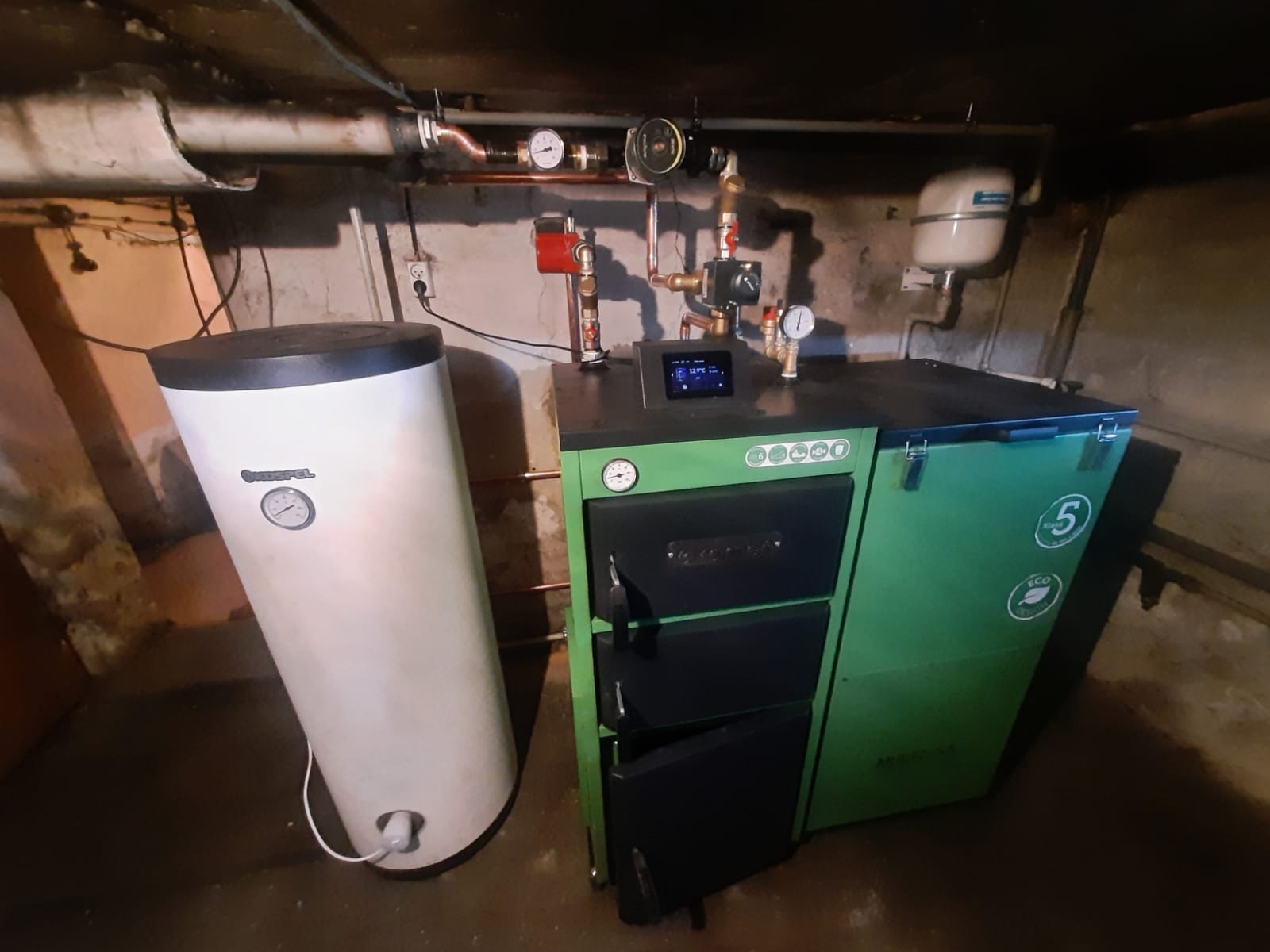 Nowoczesny, zielony piec na paliwo stałe w kotłowni, z podłączonym zasobnikiem CWU marki Kospel, widoczne rury miedziane i osprzęt instalacji grzewczej.