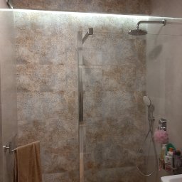 Brothers Renovation - Nowoczesna kabina prysznicowa ze szklanymi drzwiami, deszczownicą i słuchawką, wykończona płytkami o wzorzystym, beżowo-brązowym deseniu, z podświetleniem LED nad kabiną i ręcznikiem na relingu.