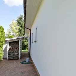 Brothers Renovation - Świeżo pomalowana elewacja budynku z widoczną fakturą tynku, brązowe rynny, skład drewna, wąż ogrodowy i wiadro na ceglanym chodniku.