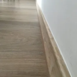 Zamontowane panele podłogowe w kolorze jasnego drewna z listwą przypodłogową przy białej ścianie, widok z niskiej perspektywy.