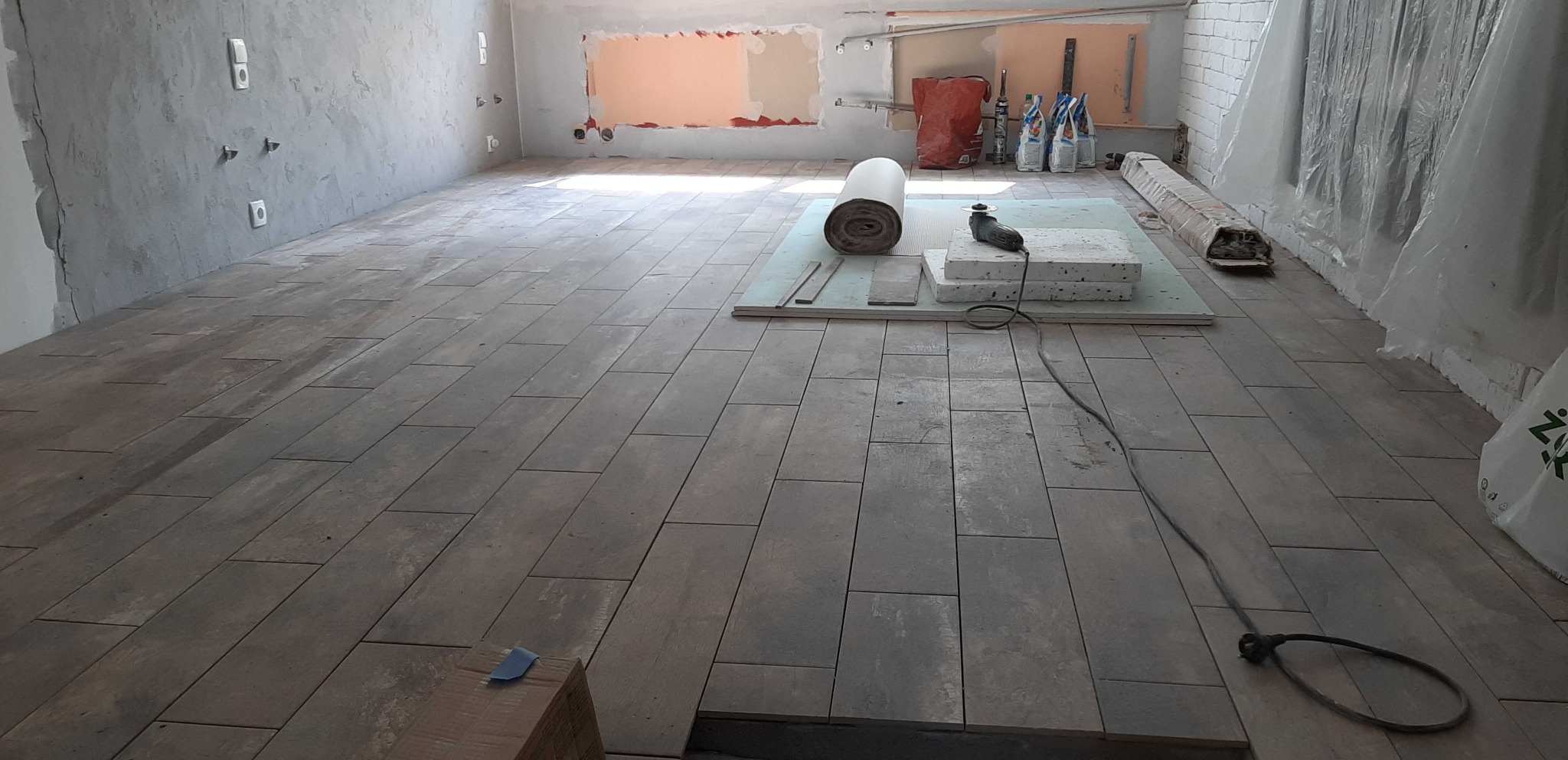 Wnętrze w trakcie remontu z ułożoną częścią podłogi z płytek imitujących drewno, narzędzia i materiały budowlane na środku pomieszczenia, ściany w stanie surowym.