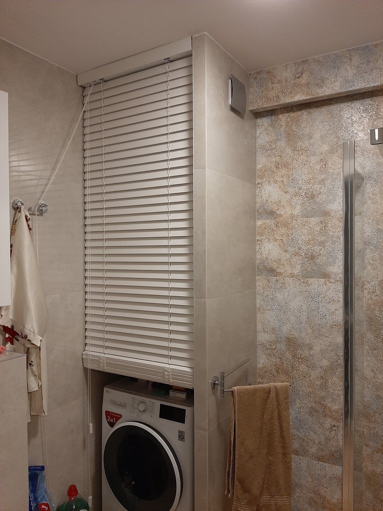 Nowoczesna łazienka z pralką ukrytą pod żaluzjami, beżowe płytki, kabina prysznicowa ze szklanymi drzwiami, ręczniki na wieszakach.