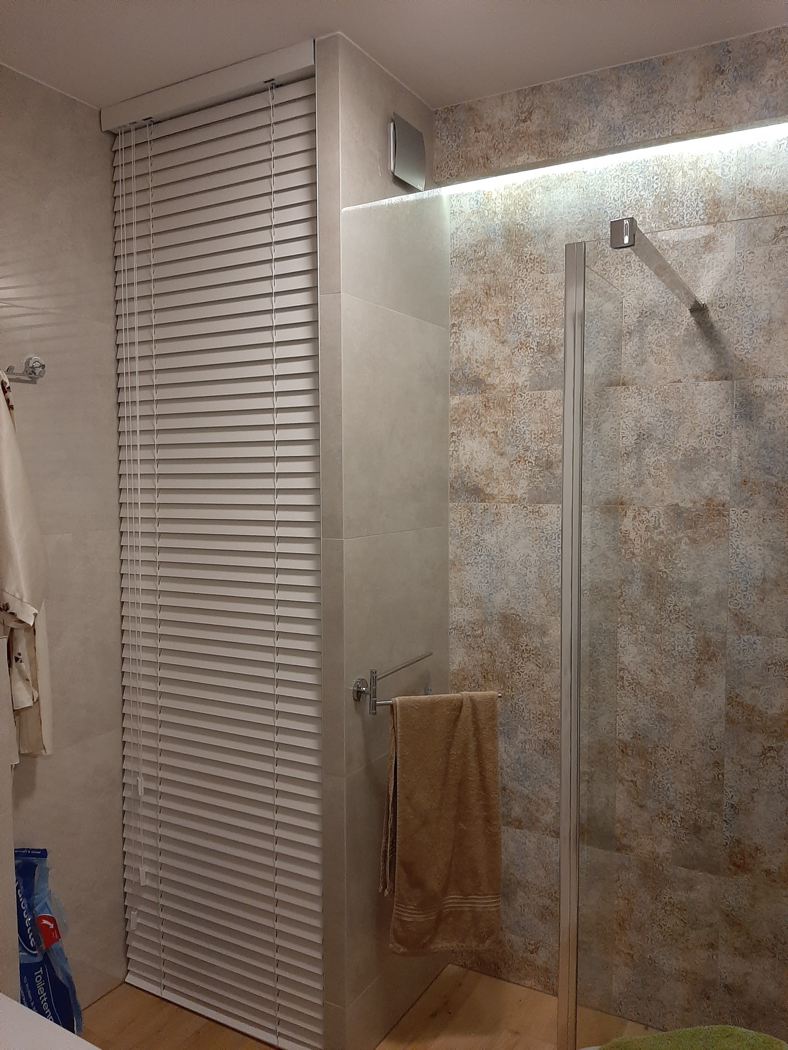 Nowoczesna łazienka z beżowymi płytkami, kabiną prysznicową ze szklanymi drzwiami, białymi żaluzjami okiennymi oraz oświetleniem LED nad kabiną.