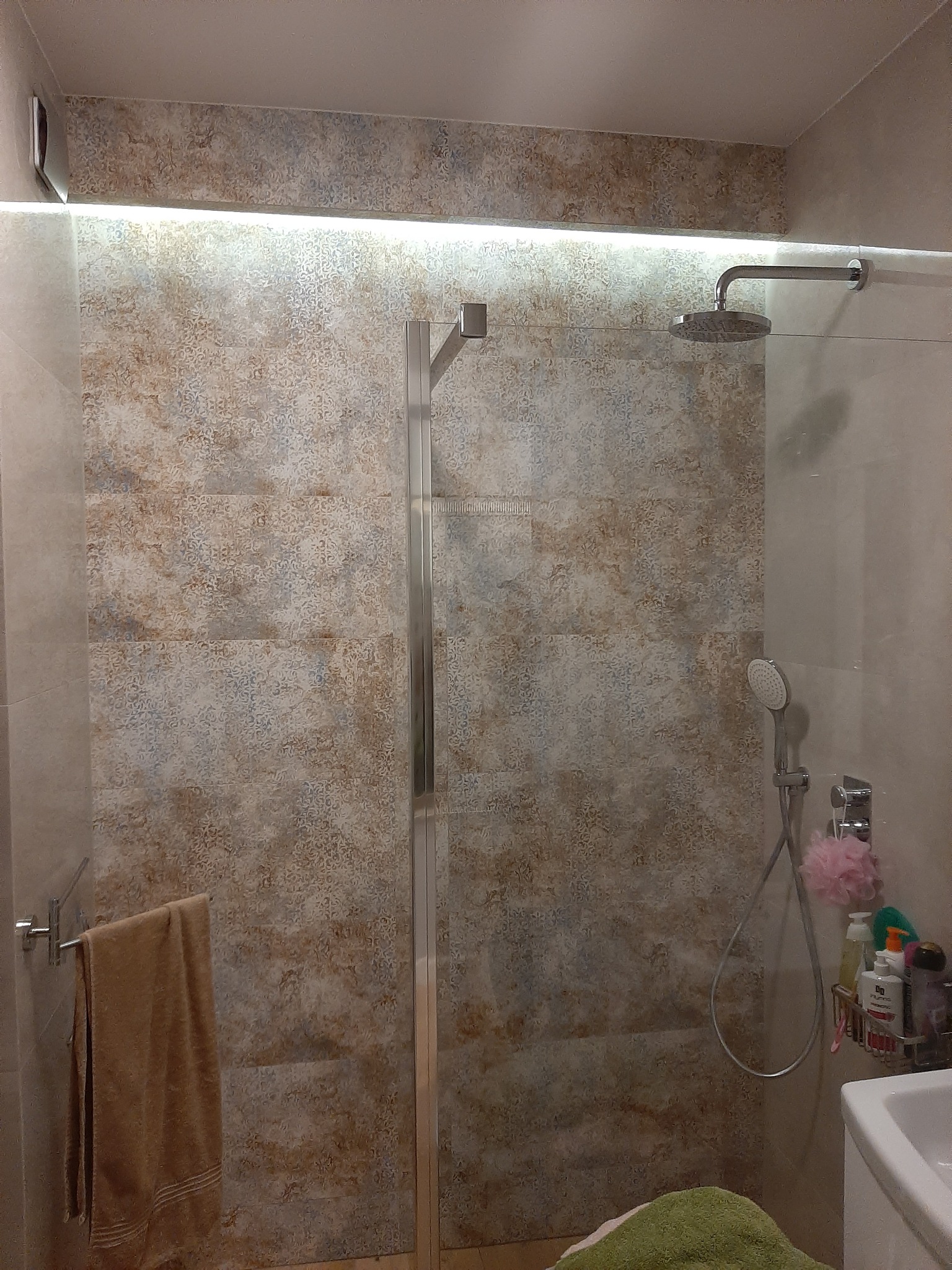 Nowoczesna kabina prysznicowa ze szklanymi drzwiami, deszczownicą i słuchawką, wykończona płytkami o wzorzystym, beżowo-brązowym deseniu, z podświetleniem LED nad kabiną i ręcznikiem na relingu.