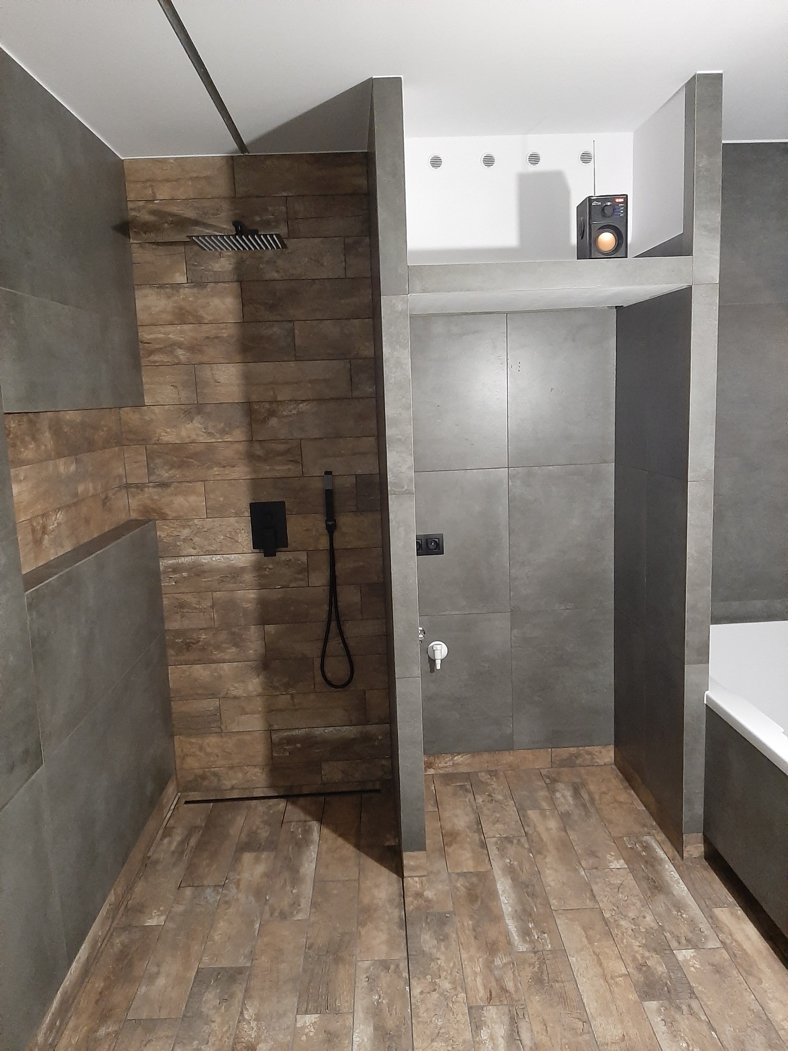 Nowoczesna łazienka z kabiną prysznicową typu walk-in wykończoną brązowymi płytkami imitującymi drewno, szara ściana z czarnym panelem sterującym prysznicem i słuchawką, oraz zabudowany głośnik...