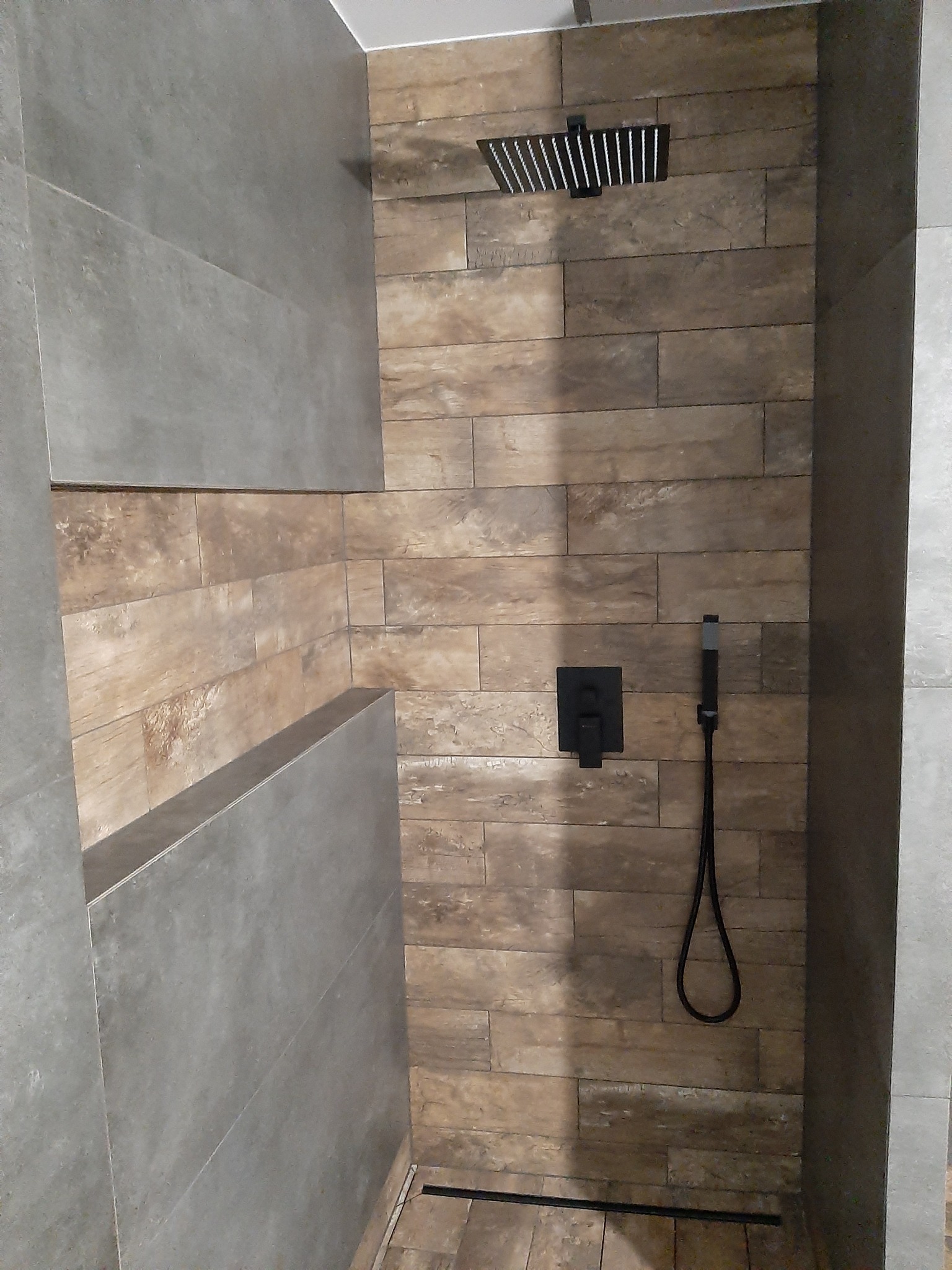 Nowoczesna kabina prysznicowa z deszczownicą i baterią w kolorze czarnym, wykończona płytkami imitującymi drewno oraz szarymi płytami betonowymi.