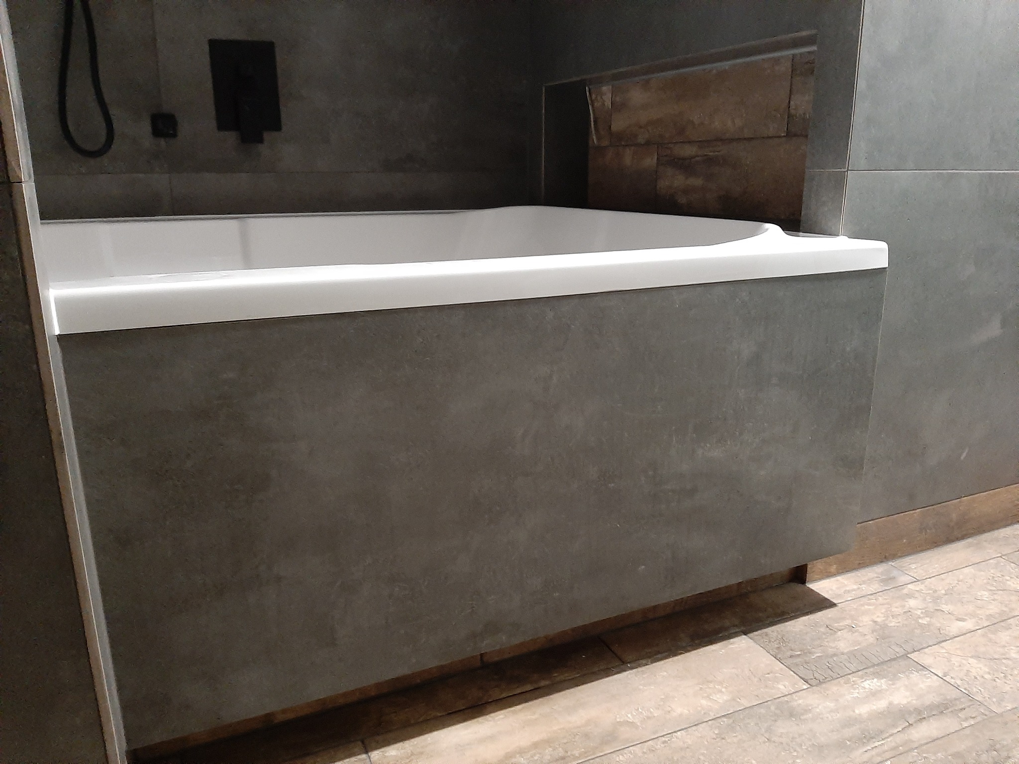 Nowoczesna zabudowa wanny w łazience z szarymi płytkami i drewnopodobną podłogą, widoczny czarny panel prysznicowy.